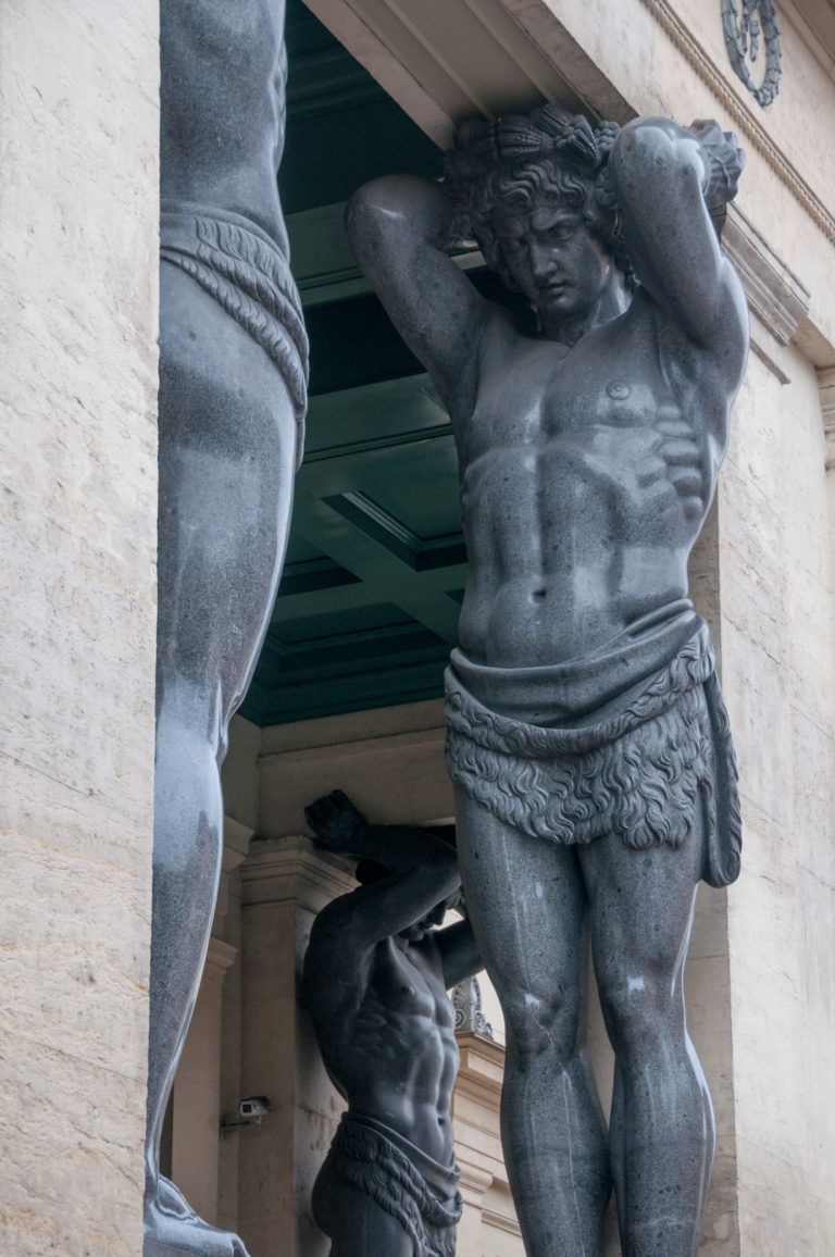Close low-angle view of a muscular dark granite Atlas figure supporting the New Hermitage portico.