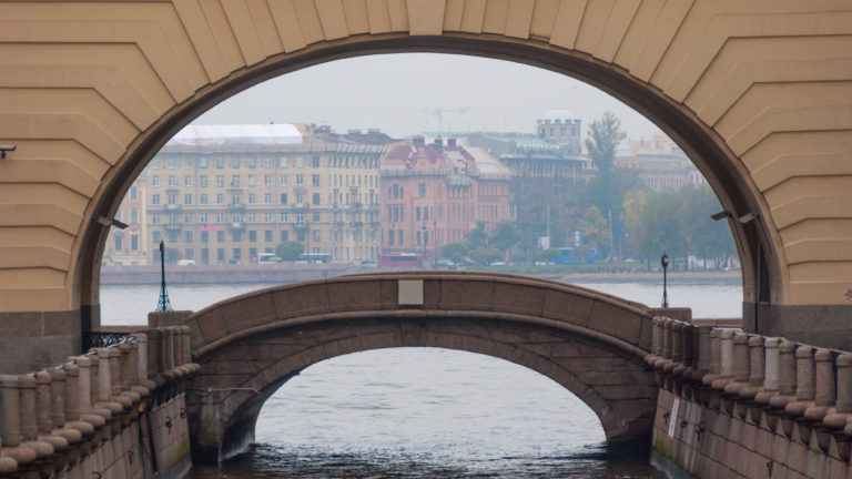 Stone arch framing a small arched bridge over the Winter Canal, with historic buildings visible beyond the water.