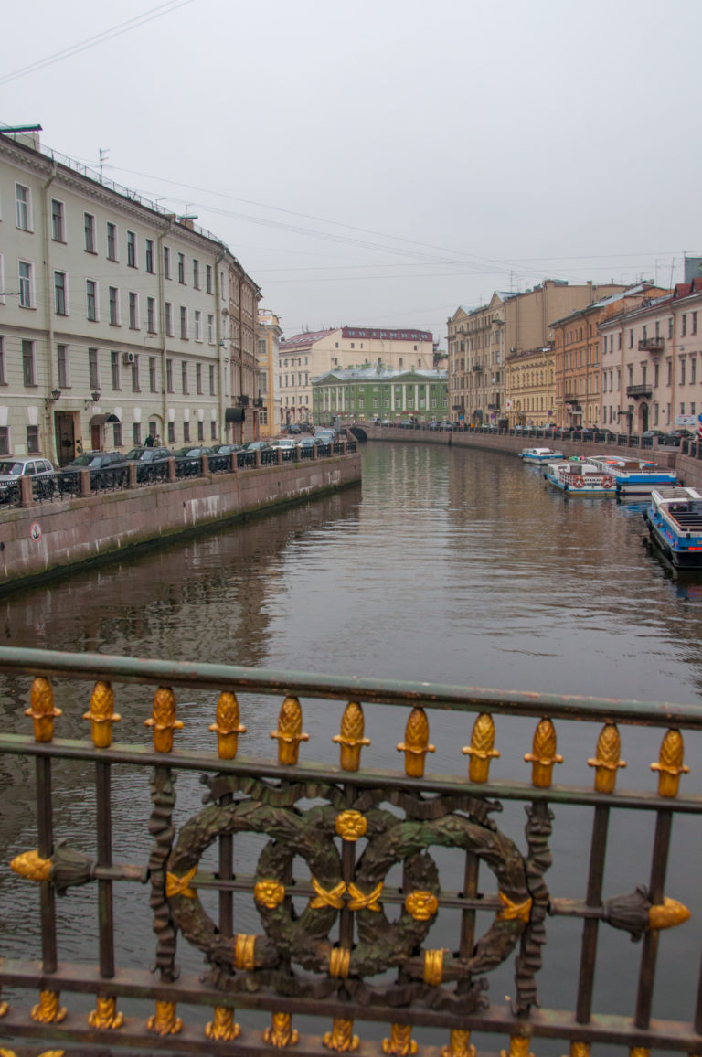 Canal bordered by historic buildings and boats, seen over a black bridge railing decorated with gilded rosettes.
