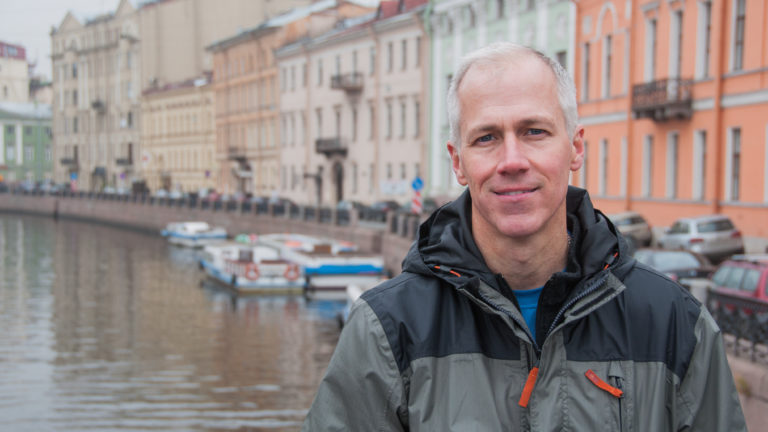 Man in a rain jacket standing beside a St. Petersburg canal with boats and pastel historic buildings behind him.