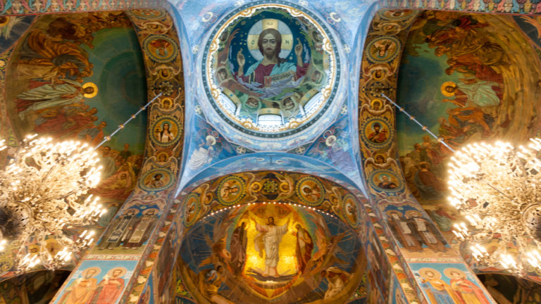 Upward view of mosaic-covered church vaults with a central Christ medallion and two large glowing chandeliers.