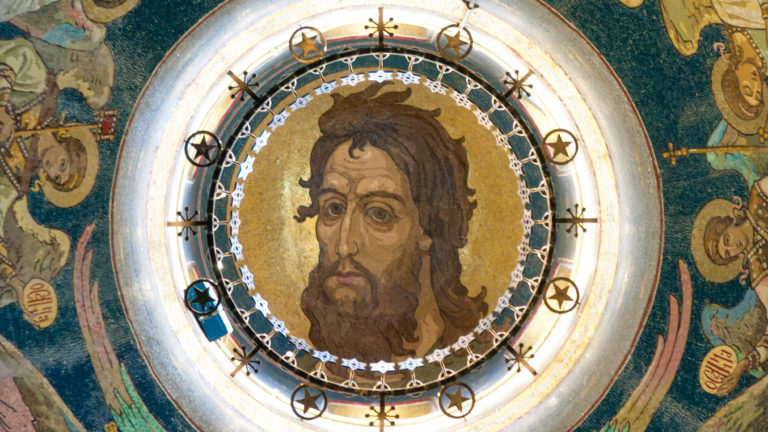 Large frontal mosaic face of Christ encircled by ornament and smaller sacred figures on a blue-green vaulted ceiling.