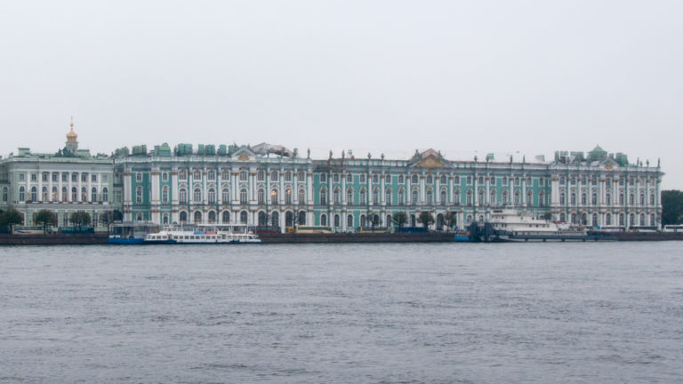 Long green-and-white Winter Palace façade viewed across the broad Neva River under a pale sky.