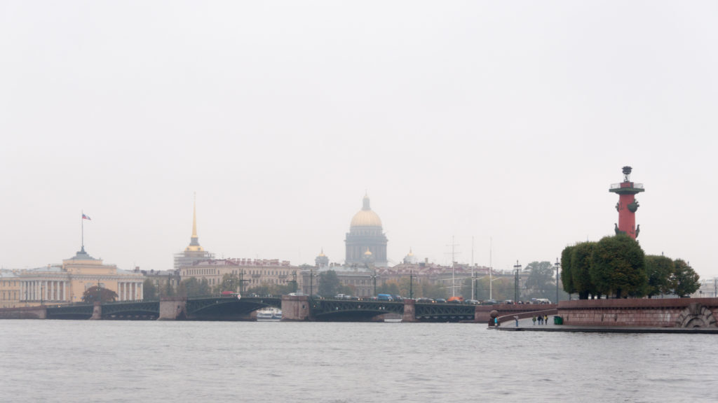 Neva River panorama with bridges, distant St. Isaac’s Cathedral dome, and a red Rostral Column at right.