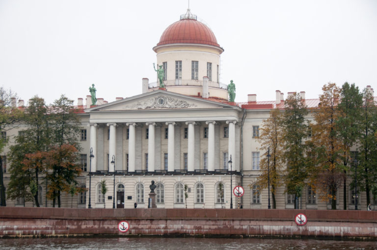 White neoclassical riverside building with a columned portico, triangular pediment, and broad reddish dome.
