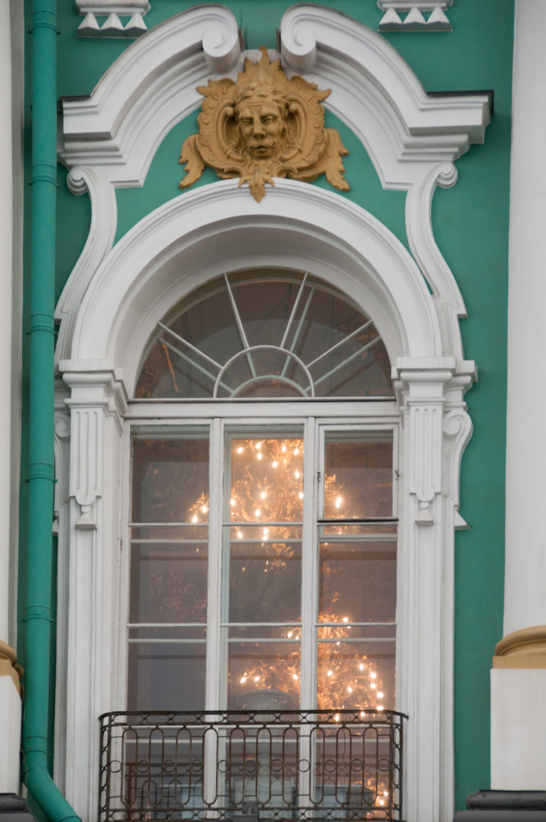 Arched Winter Palace window with white Baroque molding, gilded crest, iron balcony, and chandelier lights visible inside.
