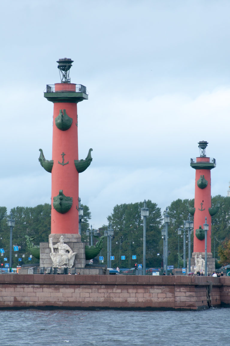 Two red Rostral Columns beside the Neva, with projecting ship prows and stone figures at their bases.