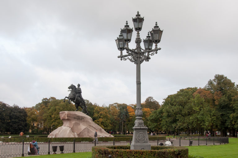 Ornate multi-lantern streetlamp in the foreground with the Bronze Horseman monument beyond autumn trees and hedges.