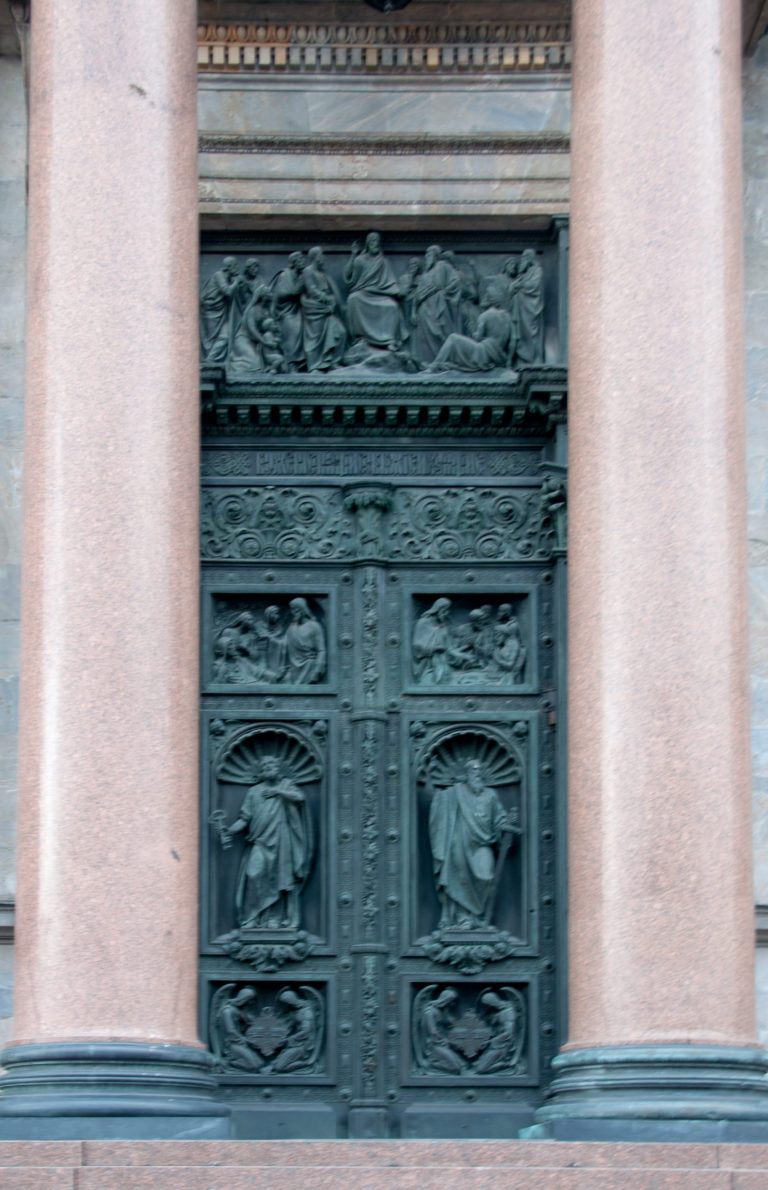 Dark patinated bronze cathedral door with multiple figural relief panels between massive polished stone columns.