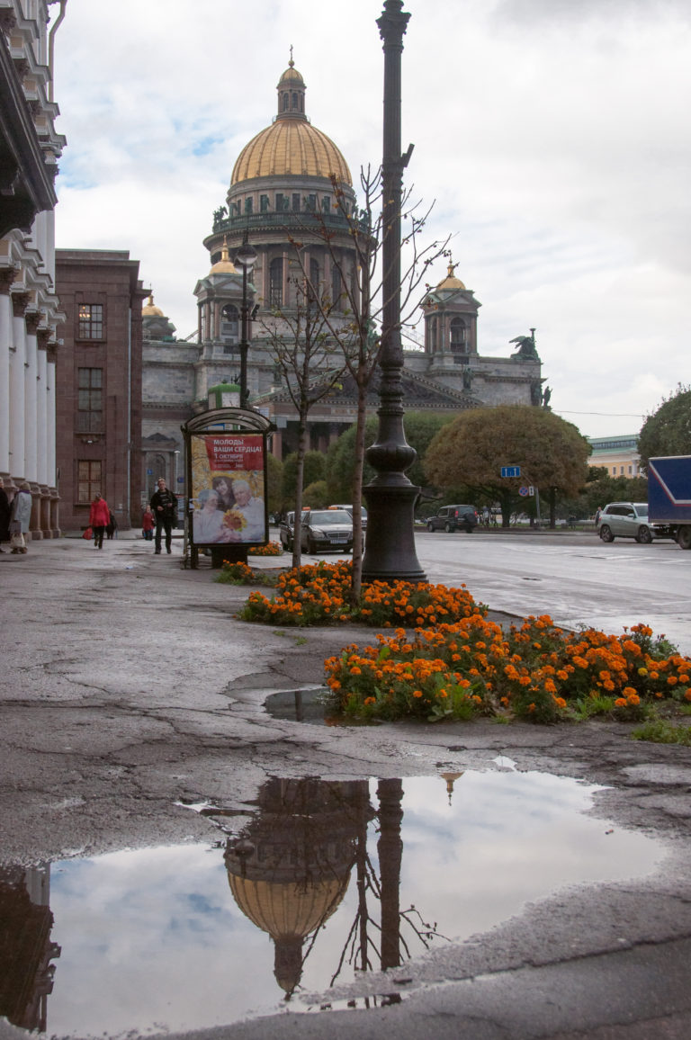 St. Isaac’s Cathedral beyond wet pavement and orange flowers, with its gilded dome reflected in a large puddle.
