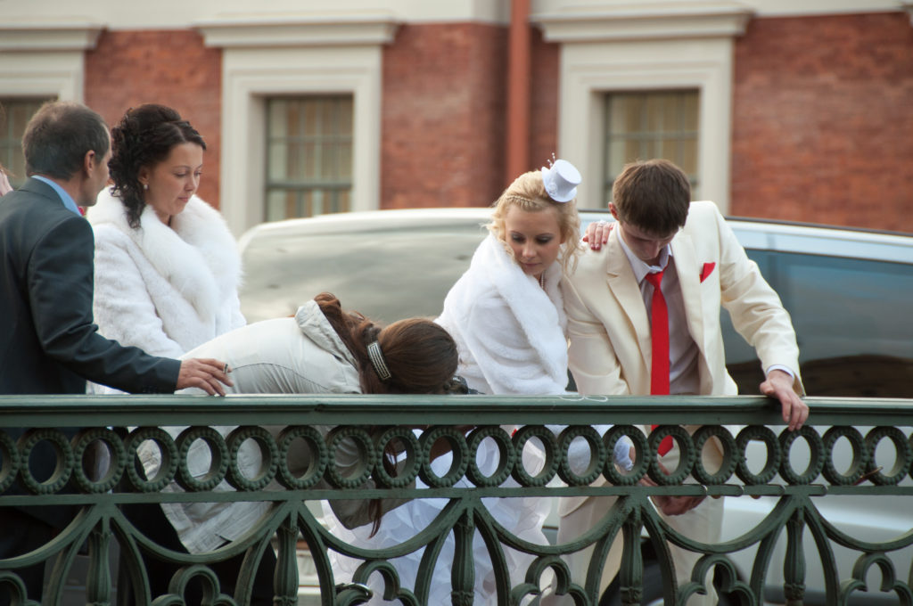 Wedding party in white formal clothing gathered beside a car and ornate bridge railing in St. Petersburg.