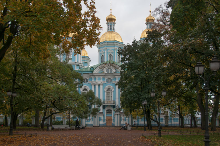 St. Nicholas Naval Cathedral seen through autumn trees across a broad path, with gilded domes above the façade.