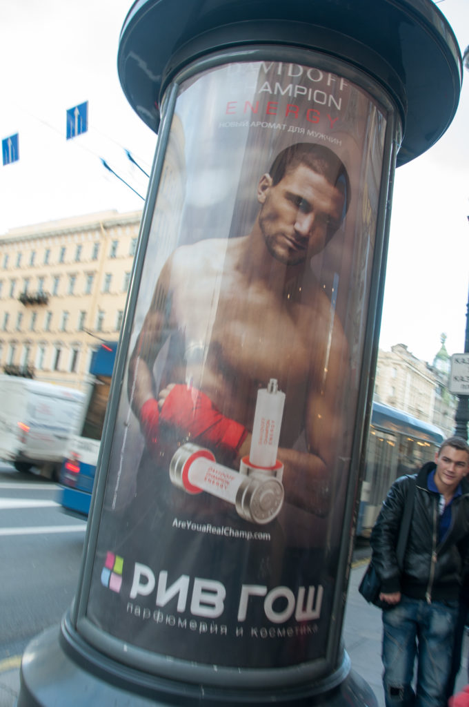 Cylindrical street advertising column with a rugby poster showing a shirtless athlete, beside traffic and pedestrians.