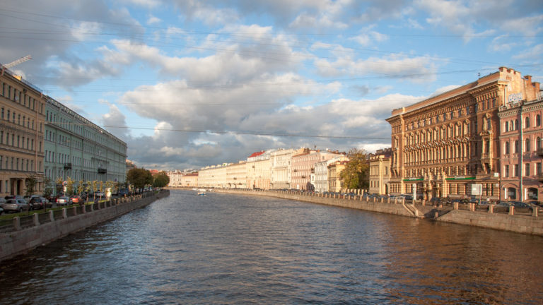 Fontanka River between rows of historic buildings, with warm sunlight on the right embankment beneath dramatic clouds.