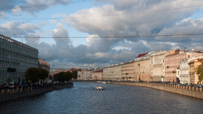 Small excursion boat on the Fontanka River between stone embankments and long rows of pale historic buildings.