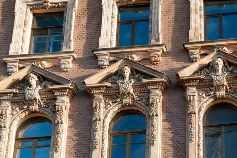 Three palace windows with elaborate pale pediments, volutes, floral carving, and female masks on a pink façade.
