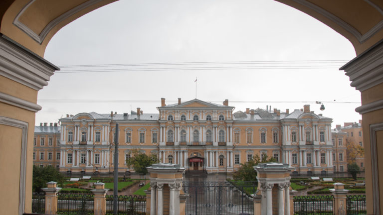 Yellow-and-white historic palace across a gated courtyard, framed by the dark curve of a large entrance arch.