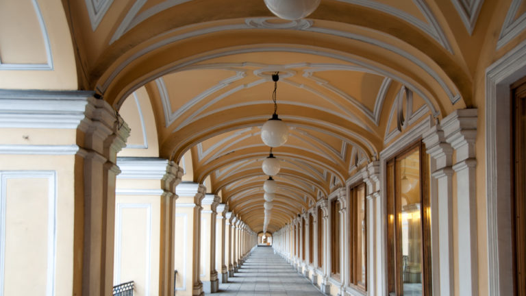 Long nearly empty ocher-and-white shopping arcade with repeated globe lights, white piers, and glass storefronts.
