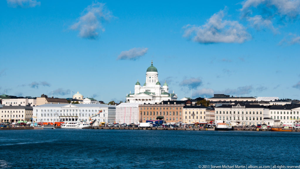 Helsinki Cathedral centered above pale waterfront buildings, viewed across blue harbor water beneath scattered white clouds.