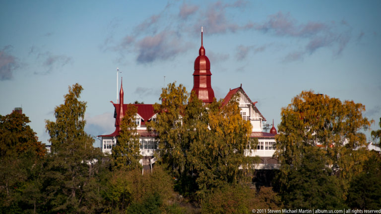 Large white historic building with a steep red roof and tall central turret rising behind yellow autumn trees.
