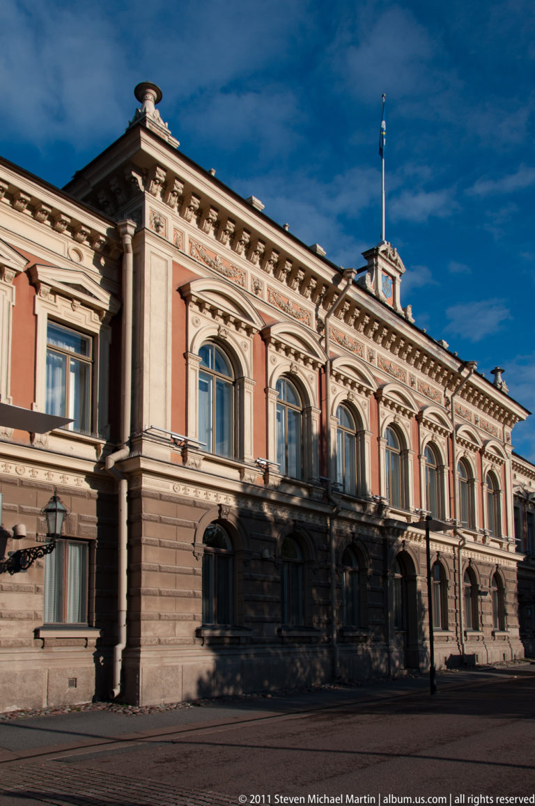 Oblique view of Ritarihuone’s ornate historic façade with arched windows, rusticated masonry, cornices, and a flagpole against blue sky.