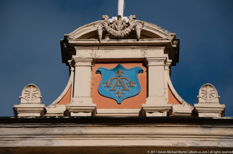 Blue shield panel with a gilded monogram framed by pale sculptural stonework and a curved pediment on Ritarihuone.