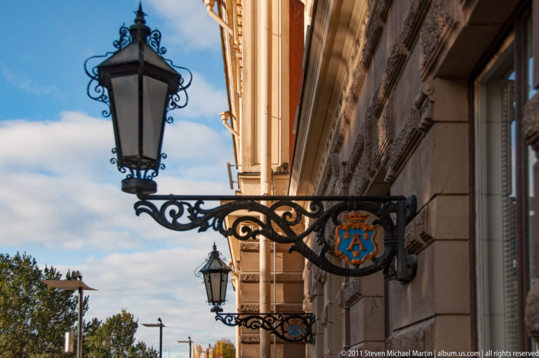 Black ornamental lantern bracket with a blue-and-orange heraldic monogram sign projecting from Ritarihuone’s stone façade.
