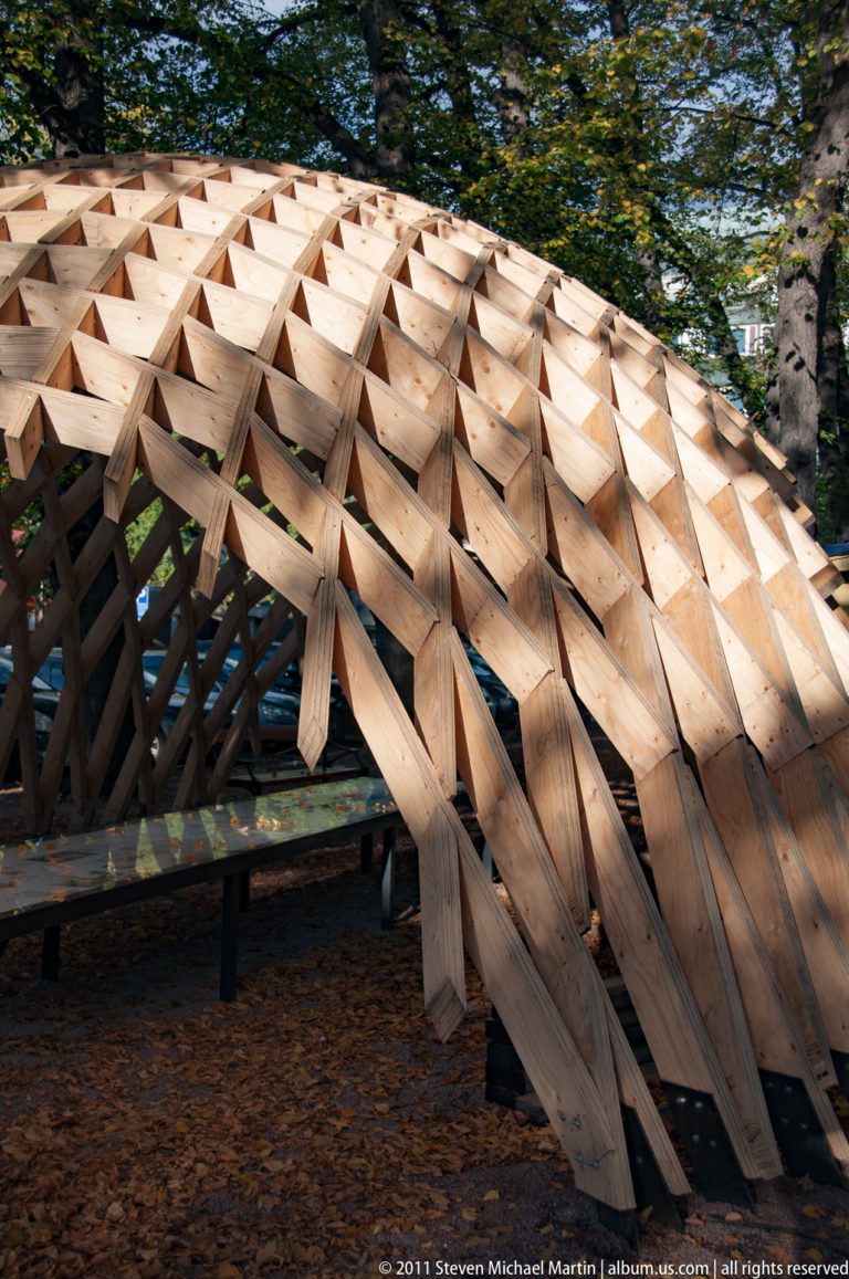 Low wave-shaped wooden lattice shelter beneath trees, with sloping timber supports and fallen autumn leaves covering the ground.