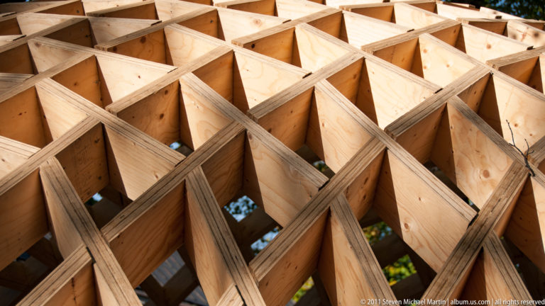 Close view of intersecting plywood ribs forming deep geometric cells, with exposed fasteners and wood grain visible.