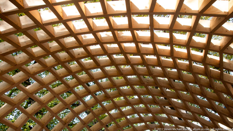 Upward view beneath a curved wooden lattice pavilion, with leaves and daylight visible through its repeating rectangular openings.