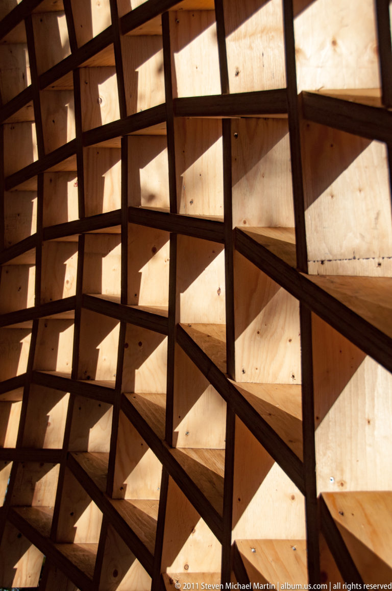 Close interior view of interlocking wooden ribs forming a curved lattice of repeating geometric cells in sunlight.