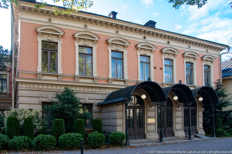 Salmon-colored historic townhouse with ornate window surrounds, clipped shrubs, and two curved black entrance canopies.