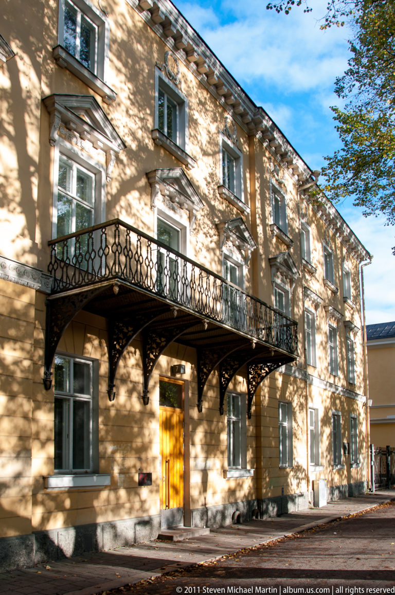 Pale ochre historic building with tall windows and a long black wrought-iron balcony casting deep shadows across the façade.