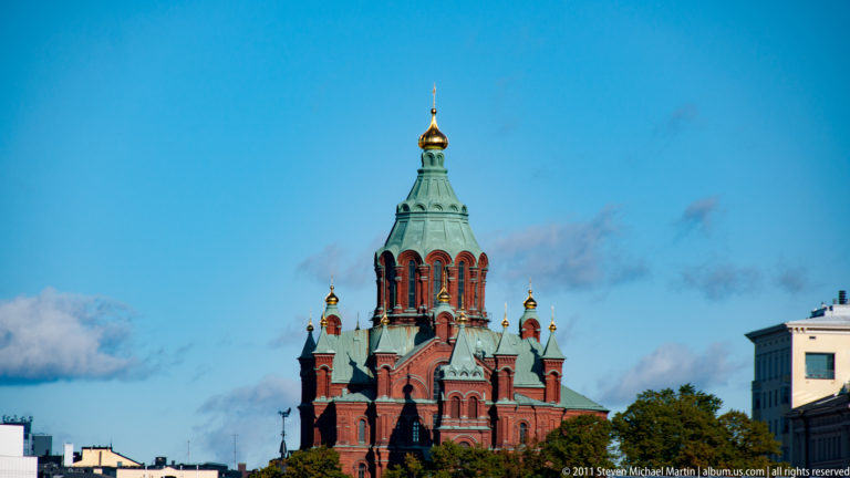 Vertical view of red-brick Uspenski Cathedral with a tall green-roofed tower and gilded onion domes against blue sky.