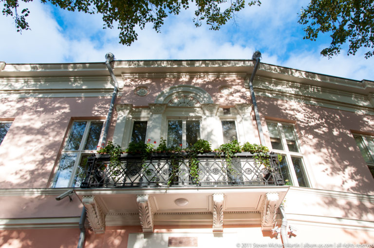 Pink stucco building façade with an iron balcony, flower boxes, white trim, carved brackets, and tree shadows.