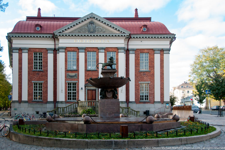Ritarihuone’s symmetrical red-brick façade behind an ornamental fountain, with bicycles and low landscaping around the forecourt.
