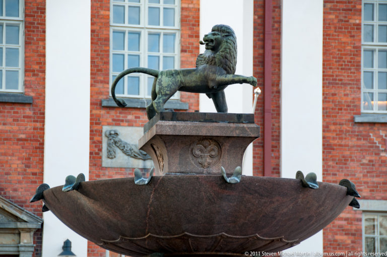 Bronze-toned lion sculpture above a broad scalloped fountain basin, with pigeons perched along the rim before a red-brick building.