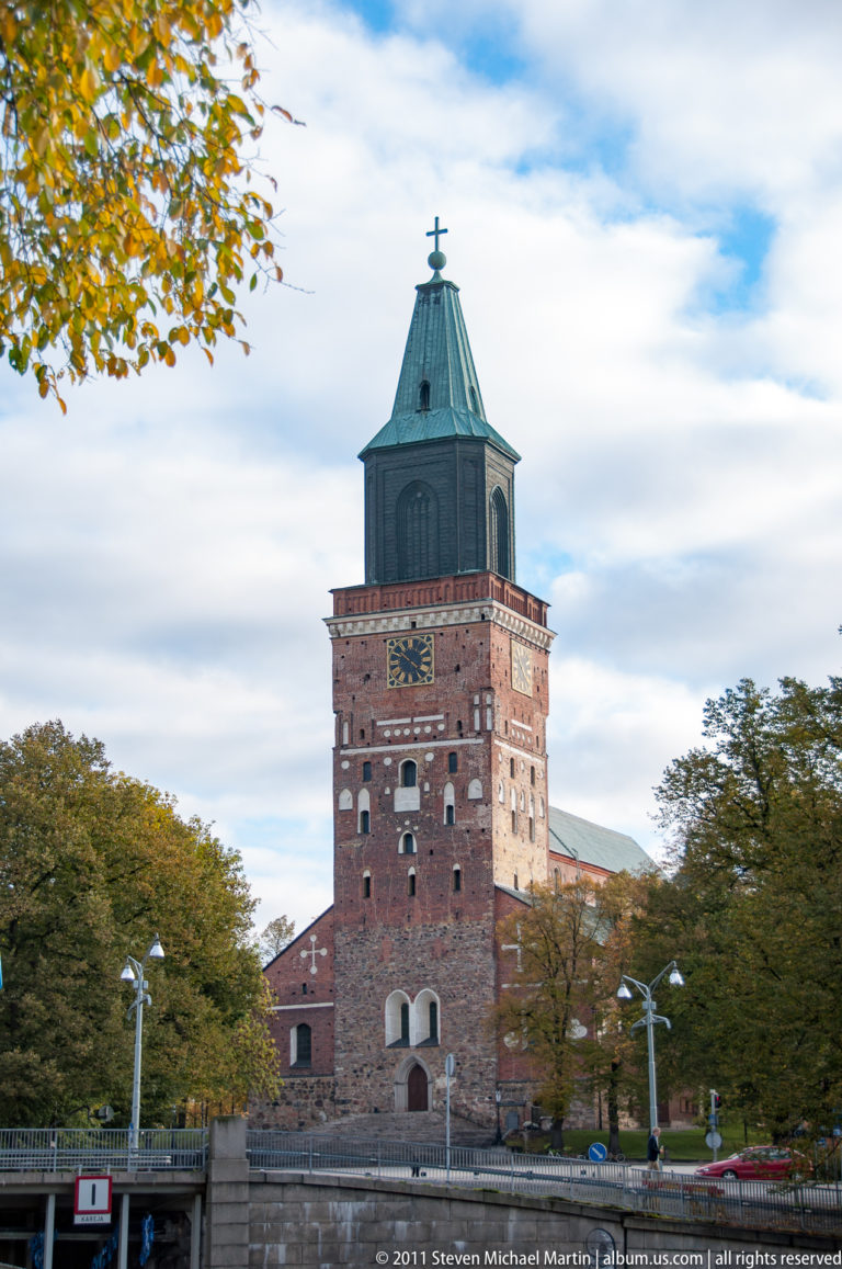 Tall historic brick tower with decorative masonry, small openings, and a green copper roof beside the Turku riverfront