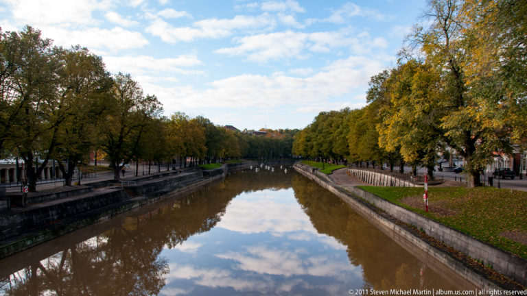 Long view down the calm Aura River between stone embankments and autumn trees, with clouds reflected in the water