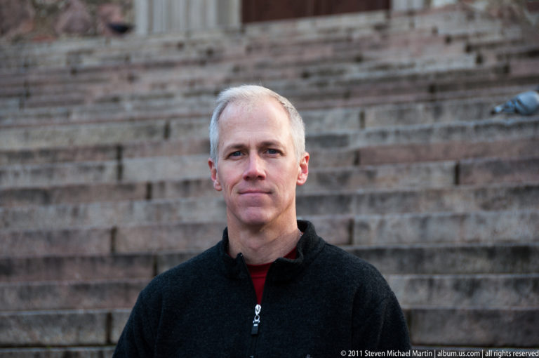 Waist-up portrait of a gray-haired man in a dark fleece standing before broad courses of weathered stone masonry
