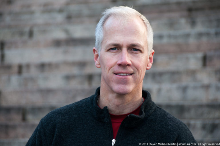 Close portrait of a gray-haired man in a dark fleece standing before a softly blurred rough stone wall