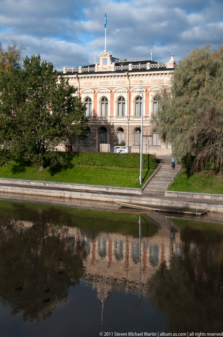 Historic riverside building with Finnish flag, trees, steps, and a clear reflection in the calm Aura River in Turku