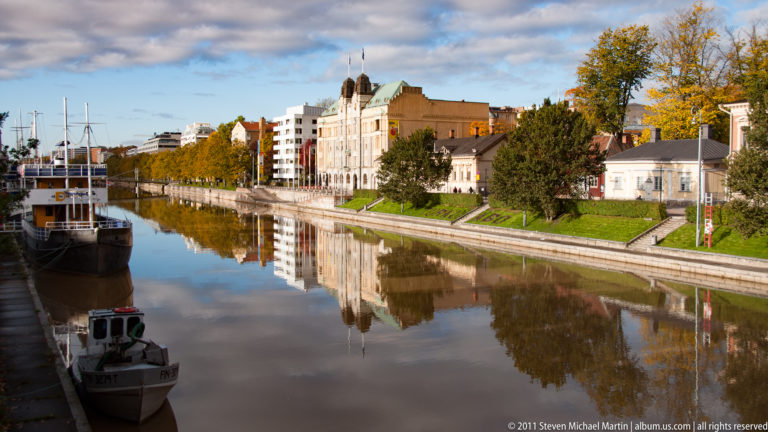 Calm Aura River in Turku with moored boats, autumn trees, embankments, and waterfront buildings reflected in the water