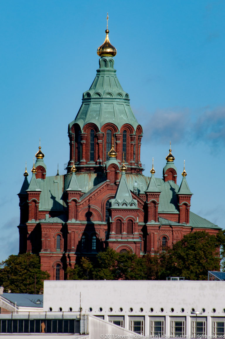 Red-brick Uspenski Cathedral with green copper roofs and gilded onion domes rising above a low white harbor foreground.