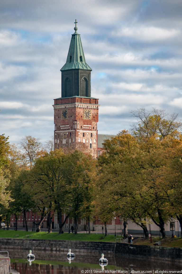 Historic brick tower with decorative bands and a green copper roof rising above trees beside the Aura River in Turku