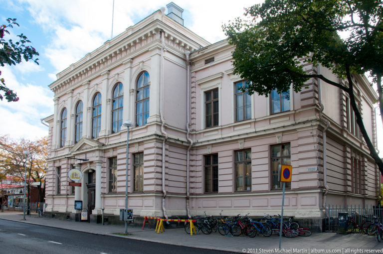Pale historic two-story building with rusticated masonry, tall arched windows, pilasters, bicycles, and trees along a city street