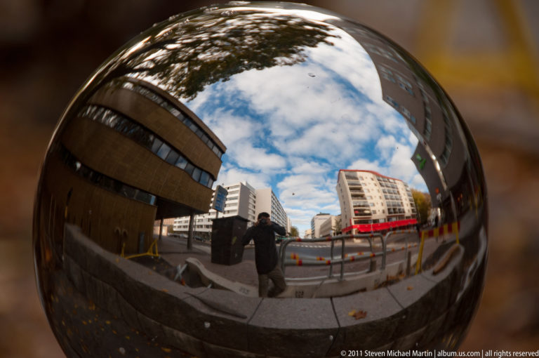 City buildings, sky, pavement, and a pedestrian distorted across the mirrored surface of a polished convex sphere