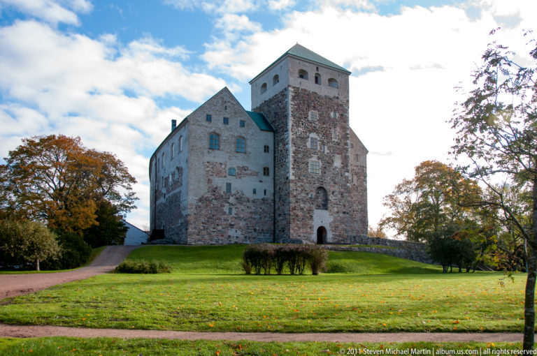 Wide view of Turku Castle beyond a green lawn and shrubs, with stone walls, steep gables, and square tower under a blue sky