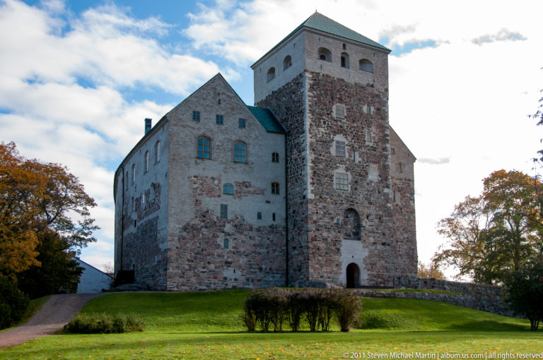 Turku Castle on a grassy rise, showing weathered stone walls, steep gables, and a tall square tower beneath broken clouds