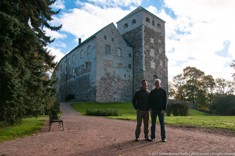 Two people standing on a gravel path before the rough stone walls, gable, and square tower of Turku Castle